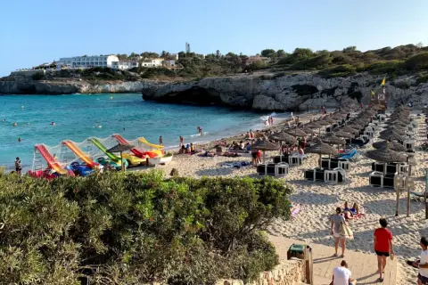 Słoneczna plaża Cala Domingos z kolorowymi zjeżdżalniami wodnymi, rzędami parasoli i turystami cieszącymi się słońcem i morzem.