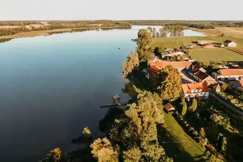 Spokojne jezioro Dejguny otoczone zielenią i domkami z czerwonymi dachami. W oddali widać lasy i pola.