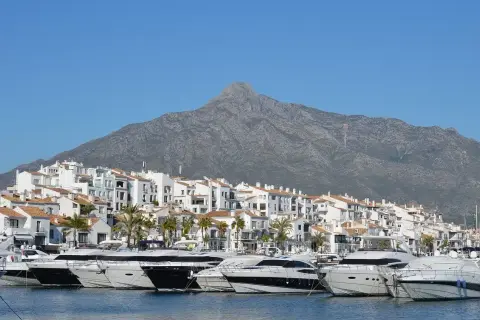 Luksusowe jachty cumują w malowniczym porcie Puerto Banús, z białymi budynkami i górą w tle.