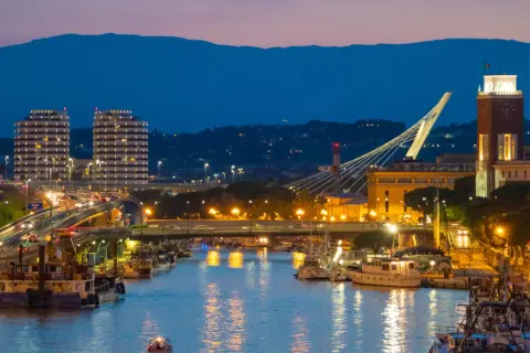 Wieczorem w Pescara: port z łodziami, nowoczesne budynki, most i wieża zegarowa.