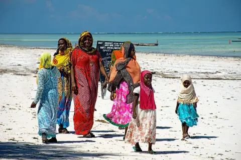 Kobiety i dzieci w kolorowych strojach spacerują po plaży Zanzibaru. Przed wyjazdem warto sprawdzić zalecenia dotyczące zanzibar szczepienia.