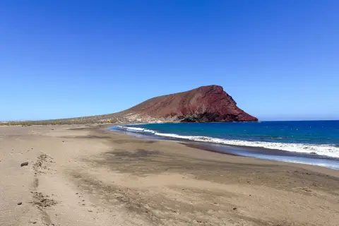 Plaże Teneryfy z charakterystyczną czerwoną górą w tle. Spokojne fale obmywają piaszczysty brzeg.