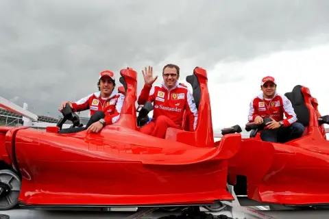Trzech mężczyzn w strojach Ferrari siedzi w czerwonym wagoniku kolejki górskiej. Jeden macha do widza.