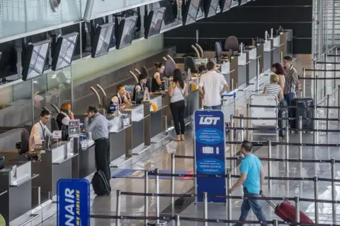 Kolejka do odprawy na lotnisku. Ludzie czekają na boarding, pracownicy obsługują stanowiska.