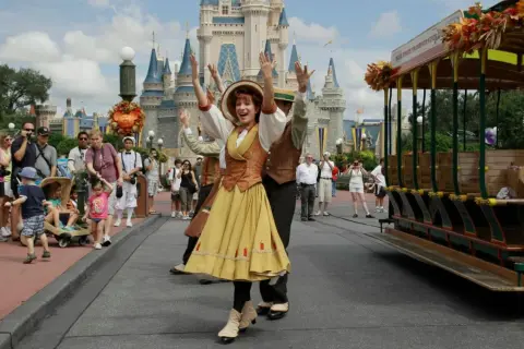 Uczestnicy parady w Disneylandzie pozdrawiają tłum. W tle zamek, a obok zabytkowy tramwaj.
