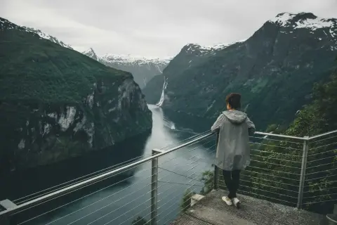 Kobieta podziwia majestatyczny fiord z wodospadem i ośnieżonymi szczytami w tle.