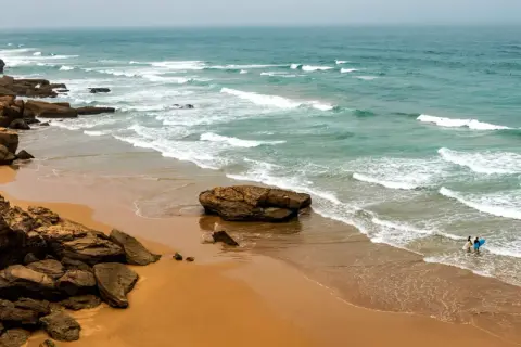 Skaliste marokańskie plaże z turkusową wodą i falami. Dwie osoby z deskami surfingowymi wchodzą do oceanu.