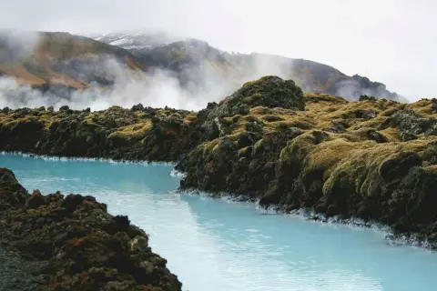 Błękitna laguna Islandia otoczona skalistym, porośniętym mchem terenem. W tle widać góry spowite mgłą i parą.