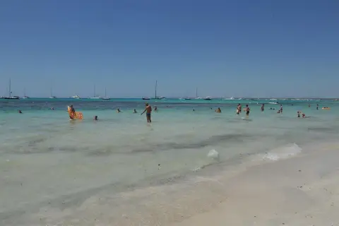 Słoneczny dzień na plaży Es Trenc. Ludzie kąpią się w turkusowej wodzie, na horyzoncie widać żaglówki.