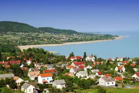 Widok na malowniczą wieś nad jeziorem Balaton. Domy z czerwonymi dachami, zielone wzgórza i błękitna woda tworzą idylliczny krajobraz. Idealne miejsce na wakacje nad Balatonem.