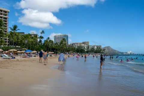 Słoneczny dzień na plaży Waikiki, z turystami kąpiącymi się w oceanie i odpoczywającymi na piasku, w tle palmy i hotele.