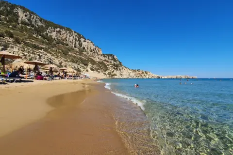 Słoneczna plaża Tsambika z piaskiem, leżakami i parasolami pod skalistym zboczem. Krystalicznie czysta woda zaprasza do kąpieli.