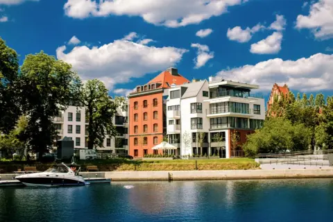 Nowoczesne budynki nad wodą, przypominające hotel Almond, z łodzią przy nabrzeżu i błękitnym niebem.