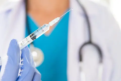 Lekarz w niebieskiej rękawiczce trzyma strzykawkę z kroplą szczepionki. Przygotowanie do szczepień.