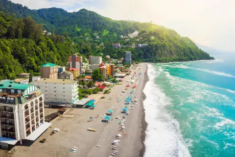 Widok na wybrzeże Gruzji, gdzie zielone wzgórza schodzą do piaszczystej plaży i turkusowego morza. Widać hotele i turystów odpoczywających nad wodą.