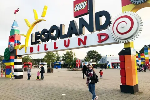 Uśmiechnięta dziewczyna z plecakiem przed wejściem do Legolandu. W tle widać bramę z napisem "LEGOLAND BILLUND" i budki z biletami.