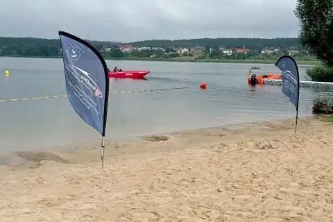 Plaża nad jeziorem, czerwona łódź ratownicza WOPR, pomarańczowa łódź, flagi, budynki na horyzoncie.