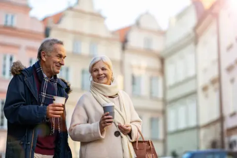 Uśmiechnięta para seniorów z kawą w rękach spaceruje po urokliwej uliczce. Marzą o podróżach, może o wycieczkach dla seniorów samolotem?