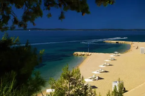 Piękna plaża przy hotelu Radisson Blu Split. Leżaki i parasole czekają na gości. W oddali widać łodzie motorowe.