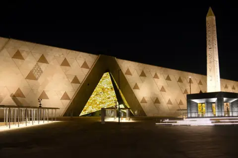 Nowe muzeum w Egipcie nocą. Monumentalna fasada z geometrycznymi wzorami i podświetlonym wejściem. Obok stoi starożytny obelisk.