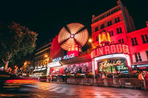Nocny Paryż rozświetlony neonami Moulin Rouge. Wirujący wiatrak i czerwone światła tworzą magiczną atmosferę.