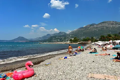 Słoneczny dzień na plażach Zatoki Kotorskiej. Ludzie odpoczywają na kamienistym brzegu, a w wodzie widać dmuchanego flaminga.