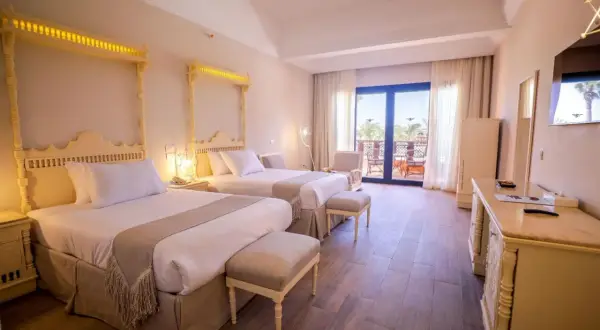 Pokój hotelowy z dwoma łóżkami, ozdobnymi zagłówkami i wyjściem na balkon. Idealne miejsce na wakacyjny calimera.
