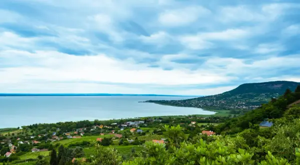 Widok na jezioro Balaton z zielonymi wzgórzami i domkami. Chmury na niebie tworzą dramatyczny krajobraz.