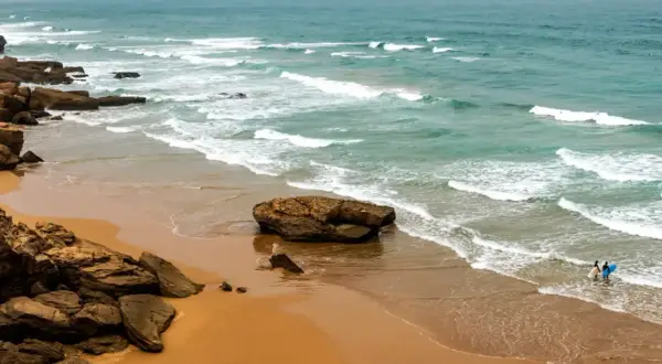 Skaliste marokańskie plaże z turkusową wodą i falami. Dwie osoby z deskami surfingowymi wchodzą do oceanu.