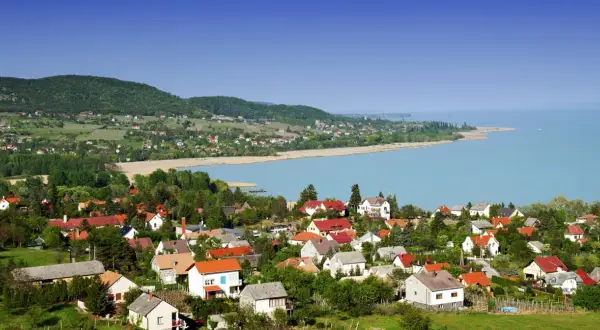 Widok na malowniczą wieś nad jeziorem Balaton. Domy z czerwonymi dachami, zielone wzgórza i błękitna woda tworzą idylliczny krajobraz. Idealne miejsce na wakacje nad Balatonem.