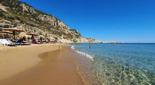 Słoneczna plaża Tsambika z piaskiem, leżakami i parasolami pod skalistym zboczem. Krystalicznie czysta woda zaprasza do kąpieli.
