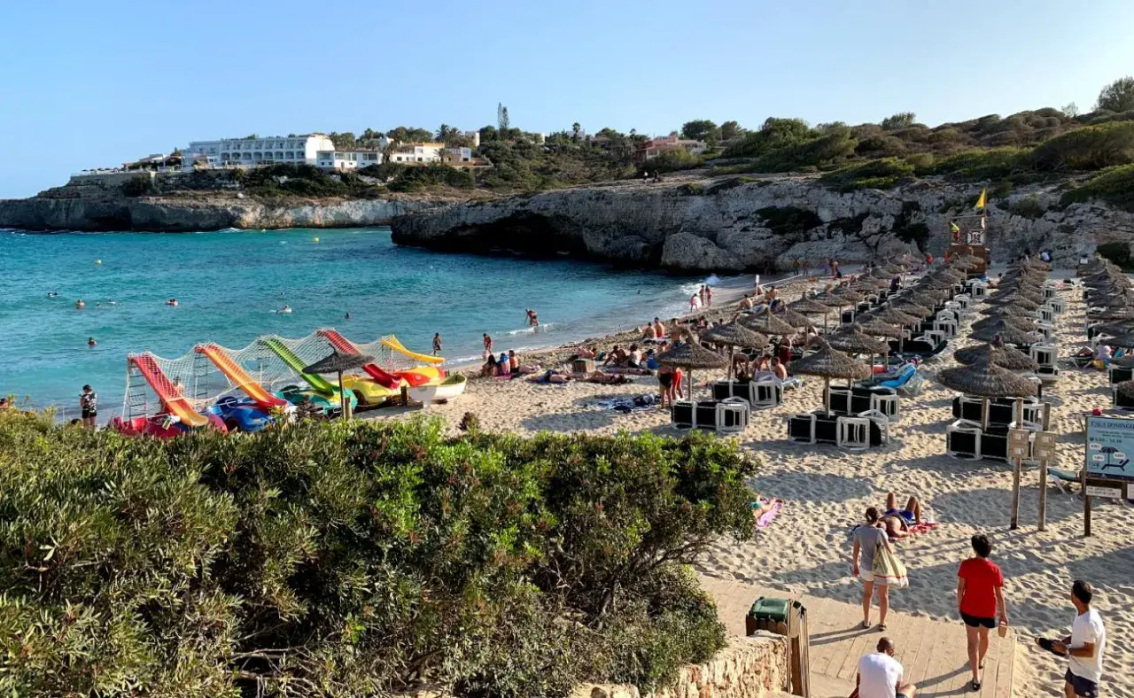 Słoneczna plaża Cala Domingos z kolorowymi zjeżdżalniami wodnymi, rzędami parasoli i turystami cieszącymi się słońcem i morzem.