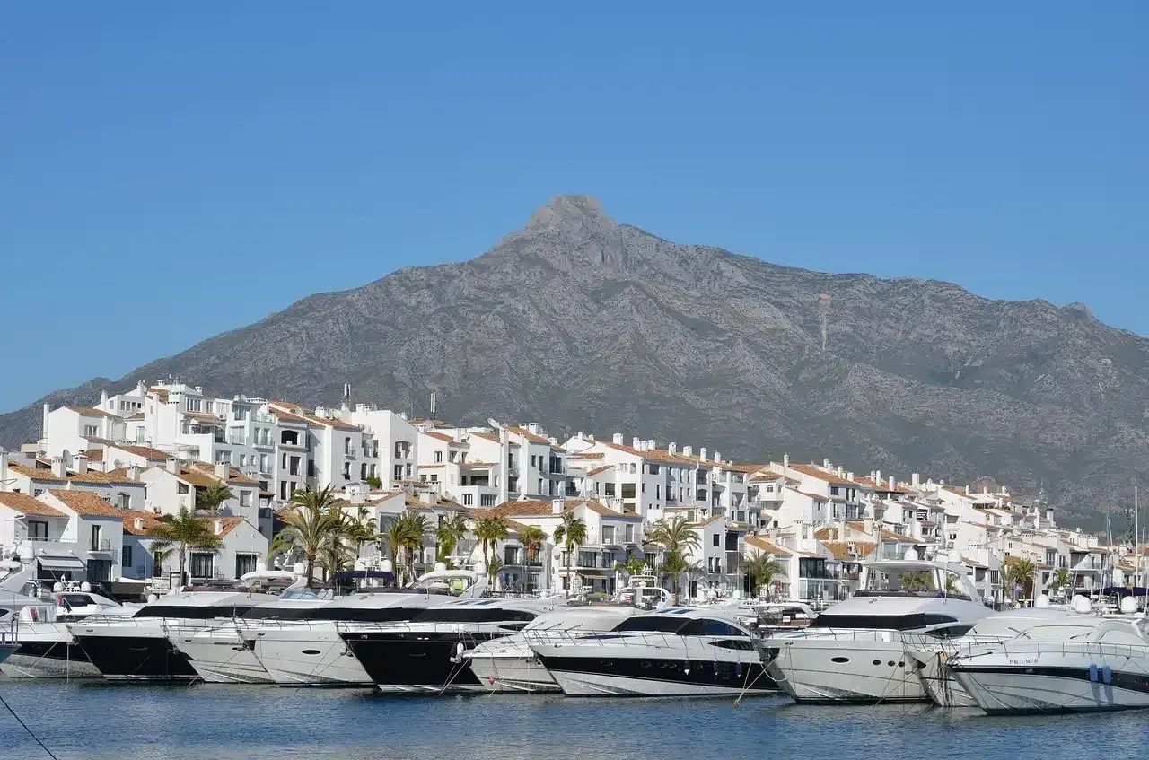 Luksusowe jachty cumują w malowniczym porcie Puerto Banús, z białymi budynkami i górą w tle.