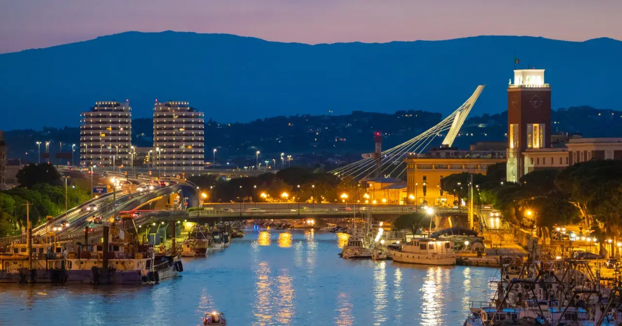 Wieczorem w Pescara: port z łodziami, nowoczesne budynki, most i wieża zegarowa.