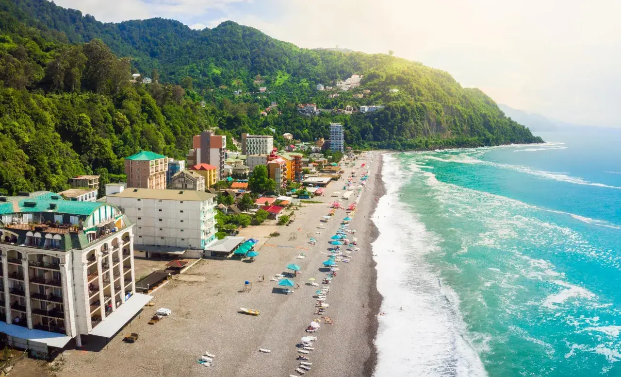 Widok na wybrzeże Gruzji, gdzie zielone wzgórza schodzą do piaszczystej plaży i turkusowego morza. Widać hotele i turystów odpoczywających nad wodą.