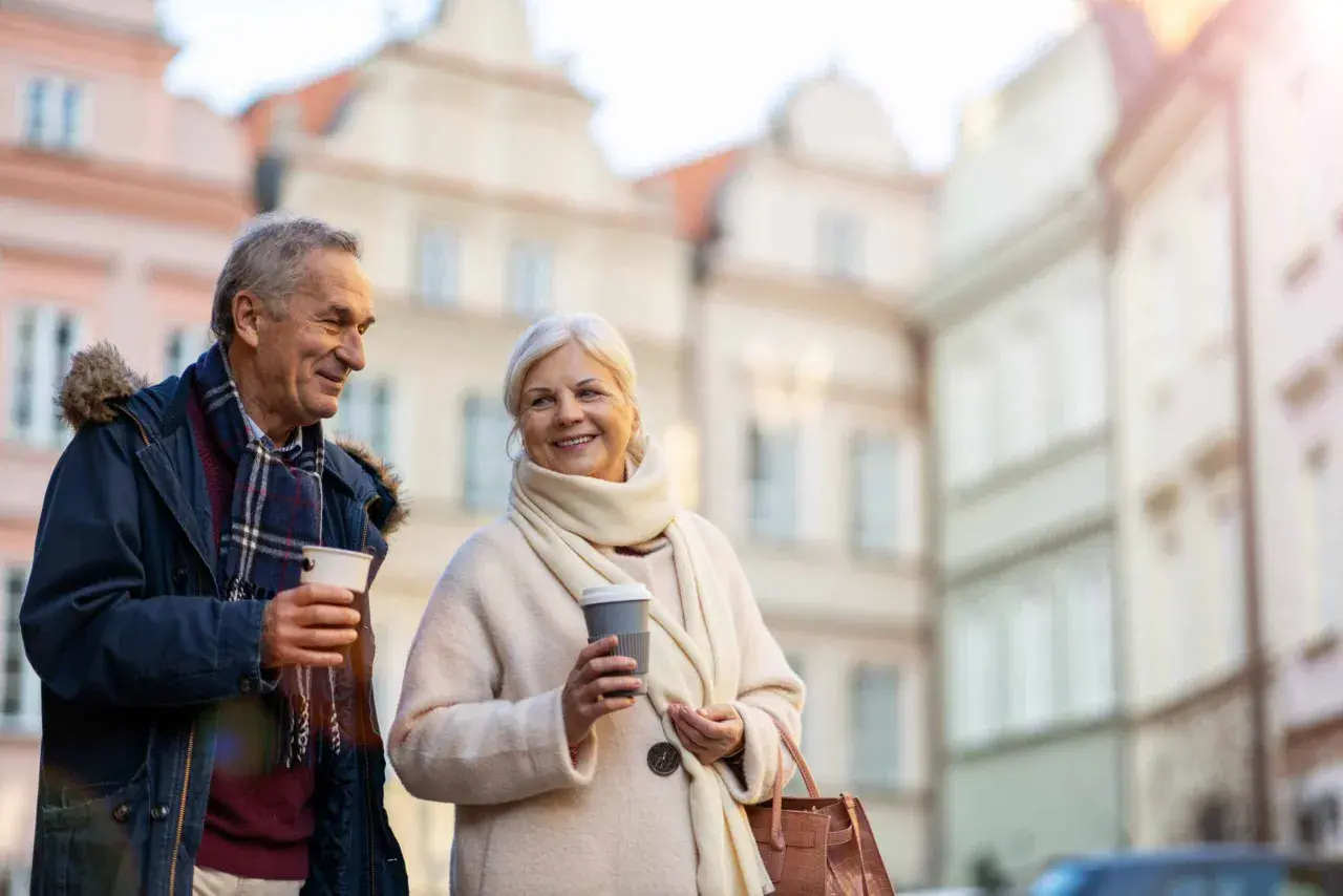 Uśmiechnięta para seniorów z kawą w rękach spaceruje po urokliwej uliczce. Marzą o podróżach, może o wycieczkach dla seniorów samolotem?