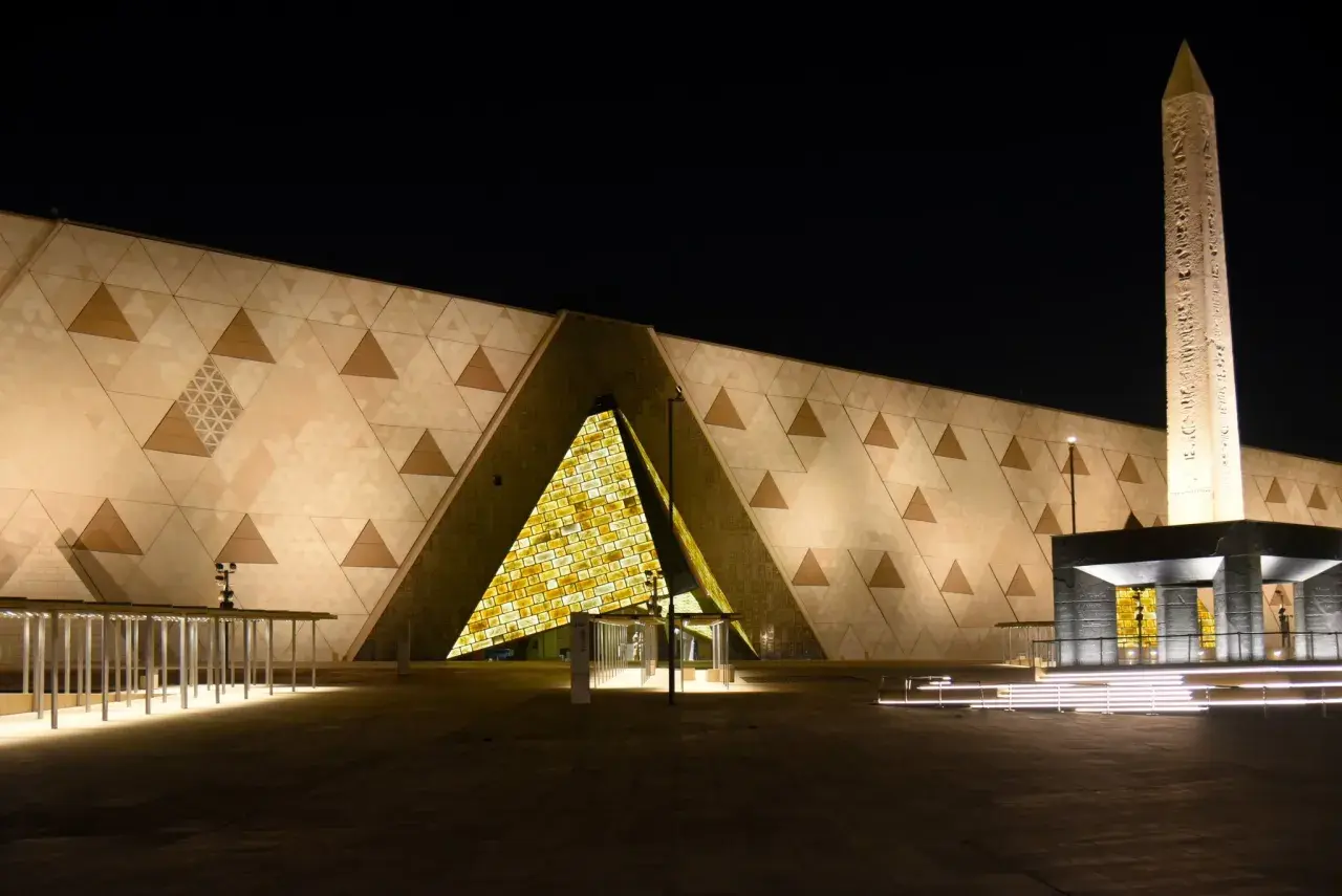Nowe muzeum w Egipcie nocą. Monumentalna fasada z geometrycznymi wzorami i podświetlonym wejściem. Obok stoi starożytny obelisk.