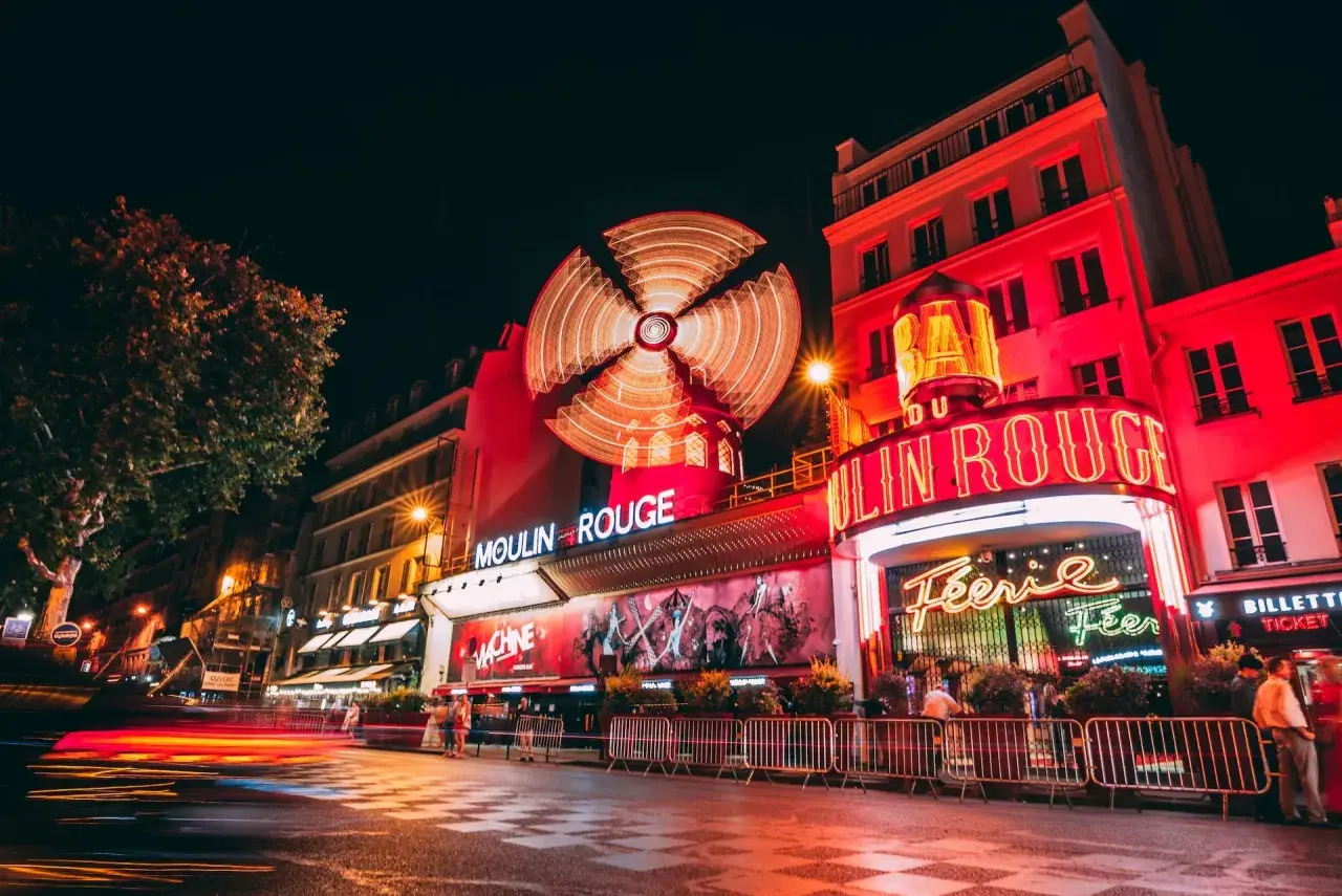 Nocny Paryż rozświetlony neonami Moulin Rouge. Wirujący wiatrak i czerwone światła tworzą magiczną atmosferę.