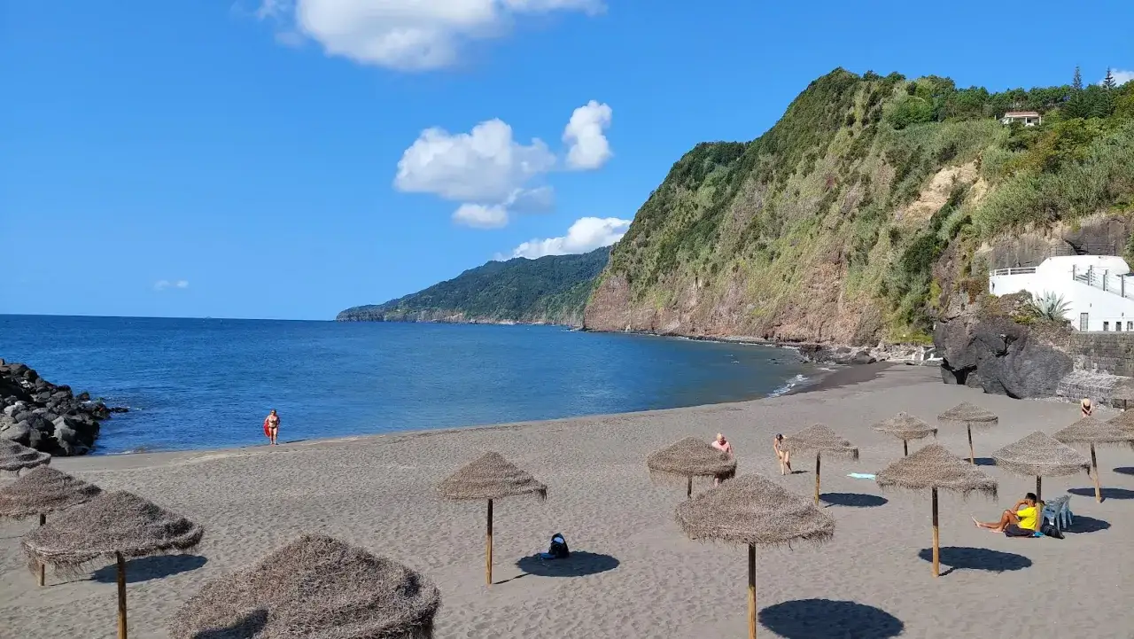 Piękne azory plaże z czarnym piaskiem, turkusową wodą i zielonymi klifami. Kilka osób odpoczywa pod słomianymi parasolami.