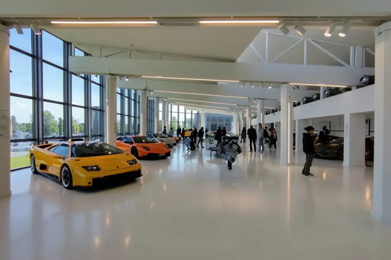 Wnętrze muzeum Lamborghini z żółtym i pomarańczowym Lamborghini, silnikiem na wystawie i zwiedzającymi.