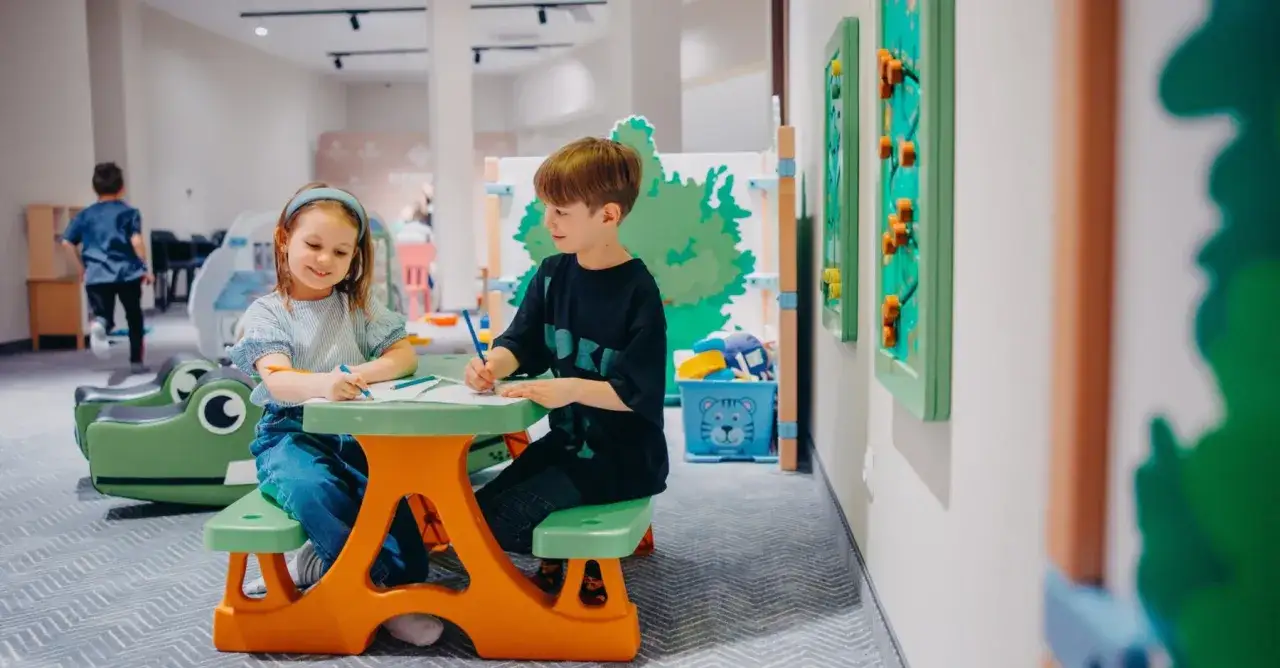 Dzieci rysują przy stoliku, a w tle widać zabawki i elementy placu zabaw. To miejsce, gdzie animator czasu wolnego dba o dobrą zabawę.