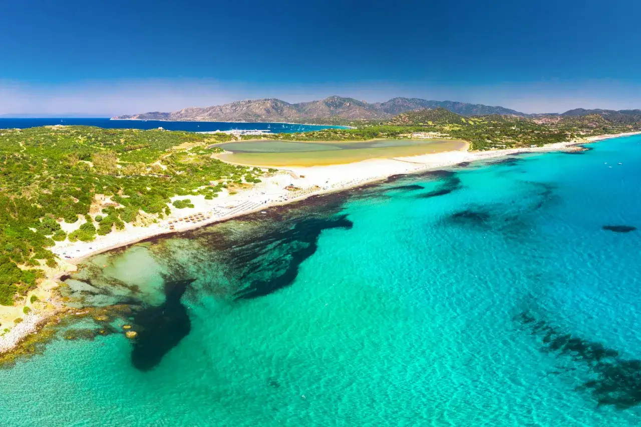 Widok z lotu ptaka na turkusowe morze i piaszczyste plaże w okolicach Cagliari. W tle góry i zatoka z łodziami.