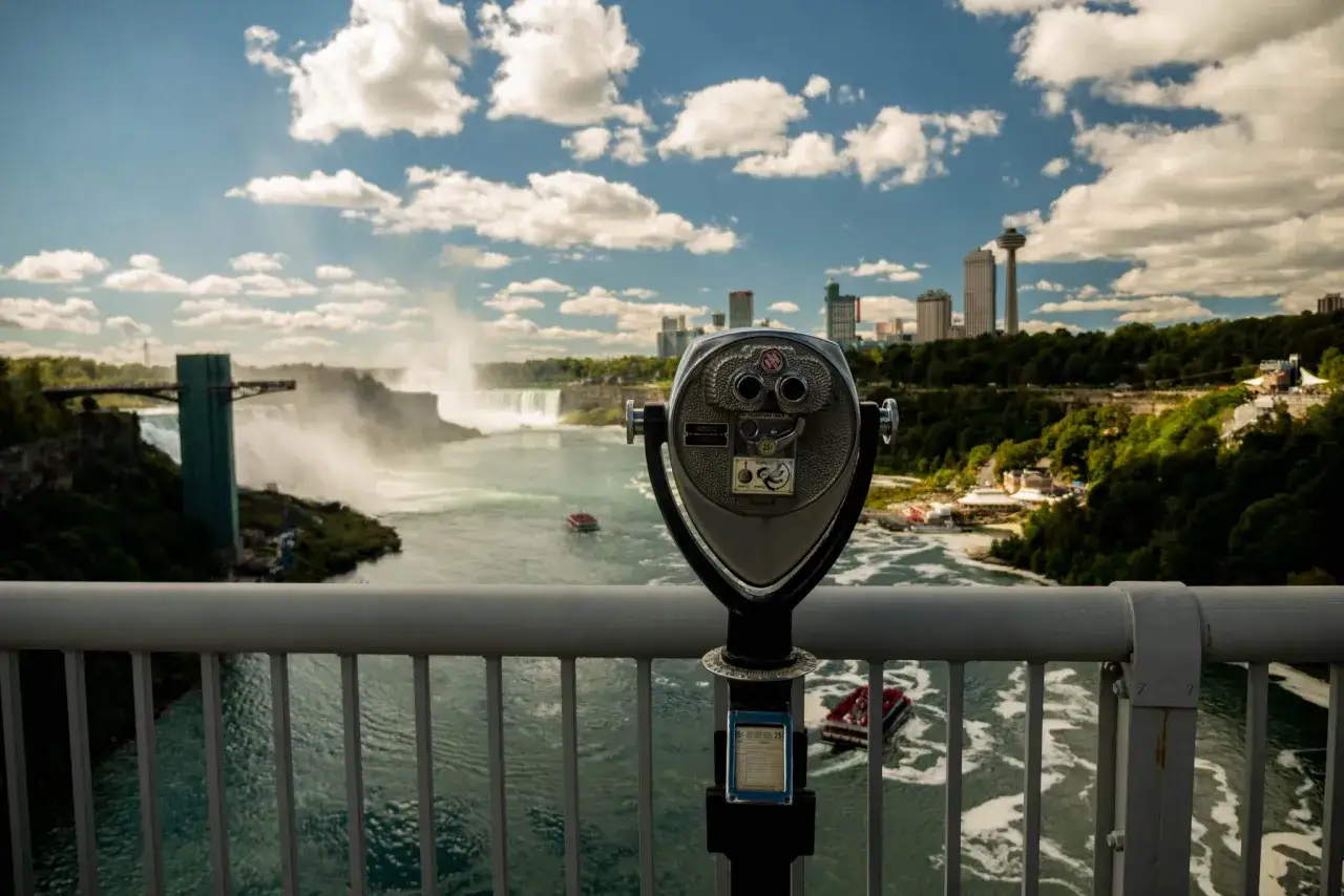 Widok na wodospad Niagara z lornetką. W oddali widać statki turystyczne i wieżowce.