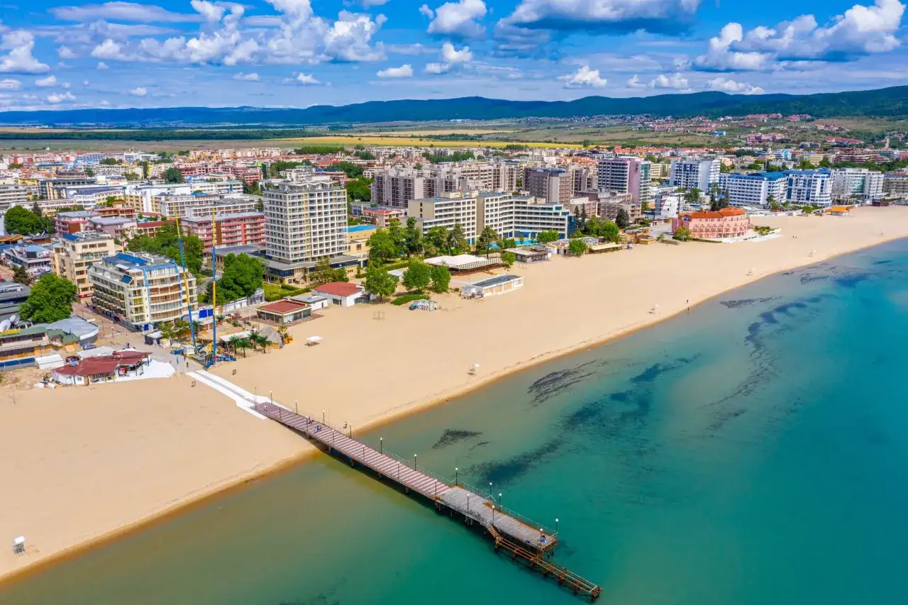 Słoneczny brzeg w pełnej krasie: szeroka, piaszczysta plaża, turkusowe morze i hotelowe zabudowania w tle. Długie molo zaprasza do spaceru.