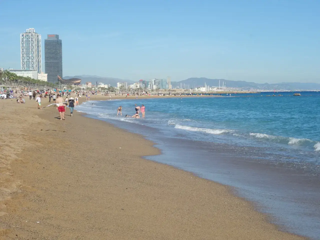 Ludzie odpoczywają na piaszczystej plaży, a w tle widać nowoczesne budynki i błękitne morze. Barcelona zaprasza do relaksu nad wodą.