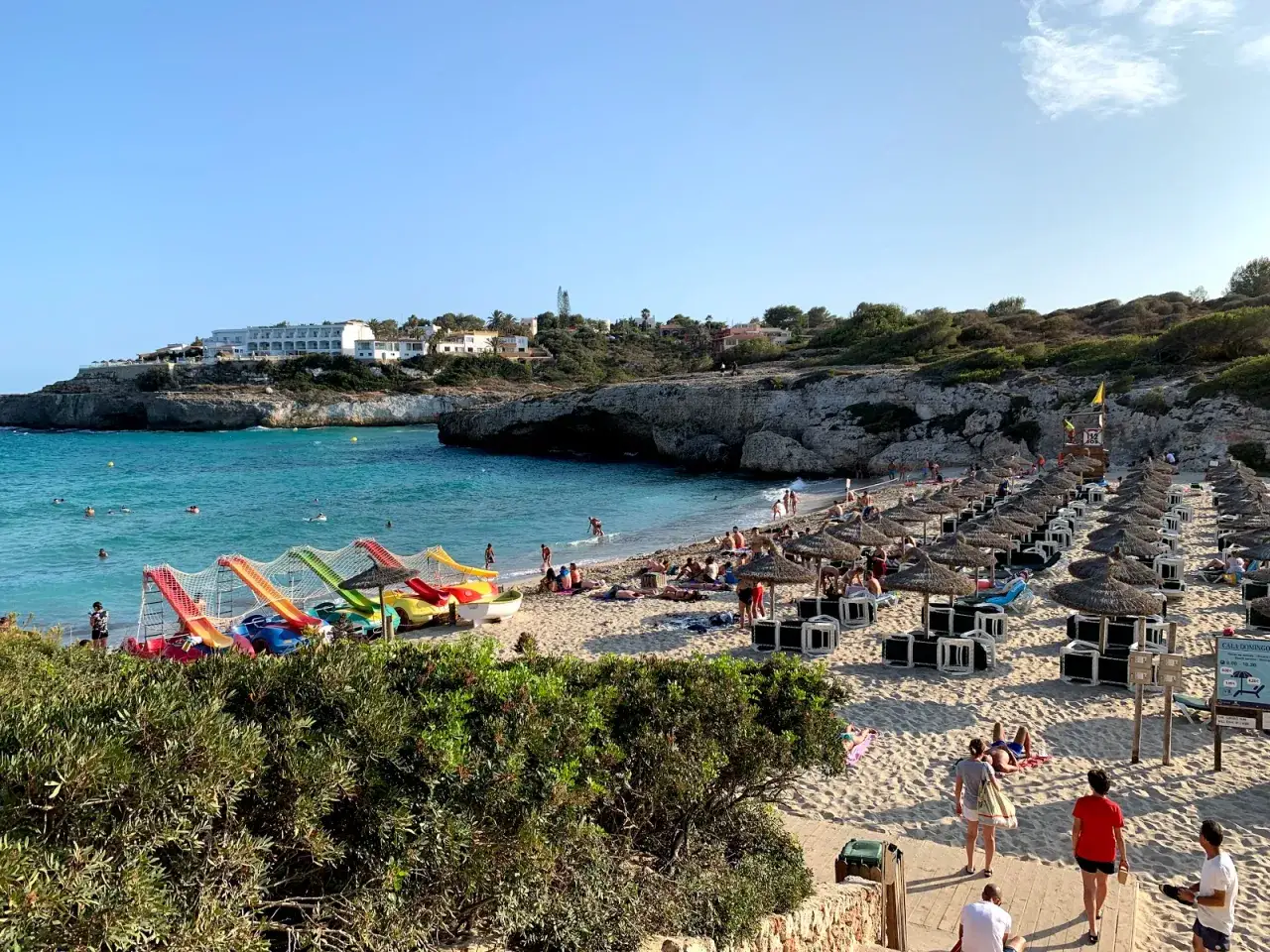 Słoneczna plaża Cala Domingos z turkusową wodą, kolorowymi zjeżdżalniami dla dzieci i rzędami parasoli.