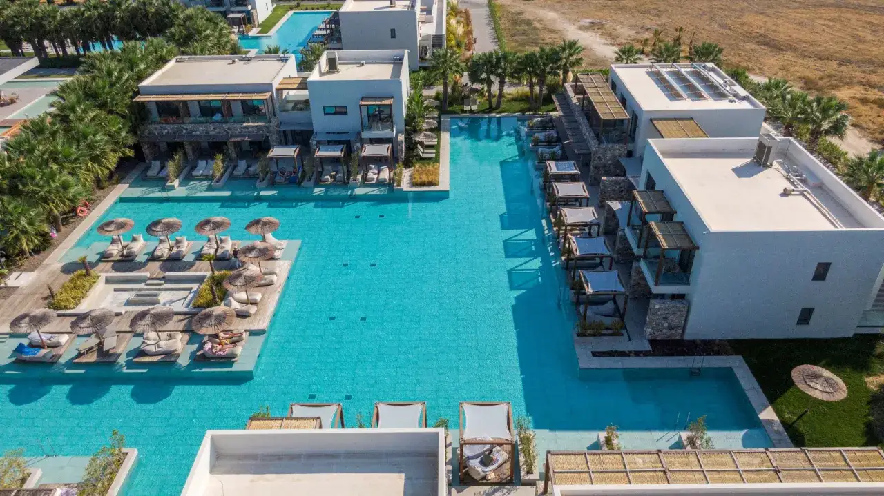 Luksusowy resort z basenami i leżakami przy plaży Caravia Beach Kos. Palmy i nowoczesna architektura tworzą idealne miejsce na relaks.