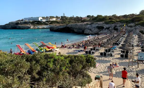 Słoneczna plaża Cala Domingos z kolorowymi zjeżdżalniami wodnymi, rzędami parasoli i turystami cieszącymi się słońcem i morzem.