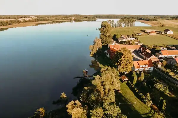 Spokojne jezioro Dejguny otoczone zielenią i domkami z czerwonymi dachami. W oddali widać lasy i pola.