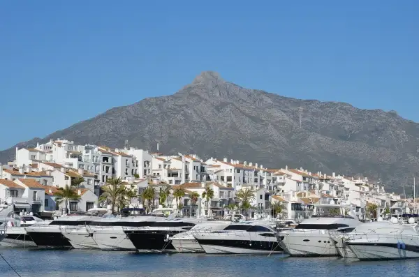 Luksusowe jachty cumują w malowniczym porcie Puerto Banús, z białymi budynkami i górą w tle.